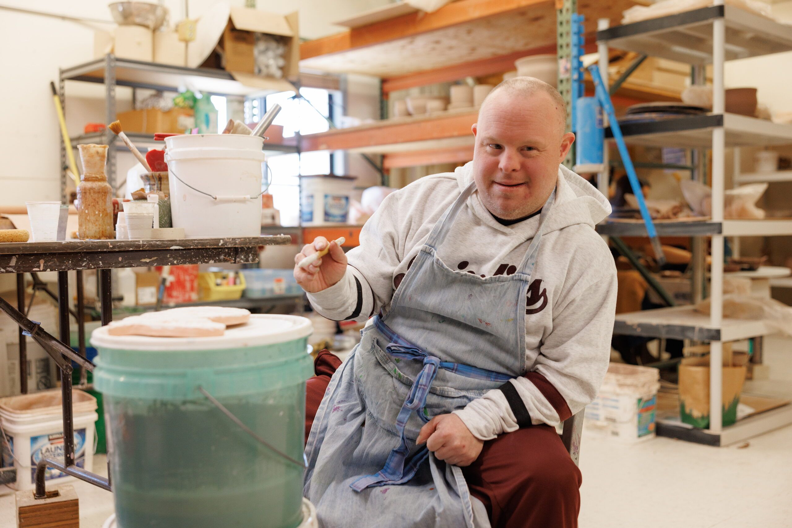 A smiling artist in a gray apron holds up a pottery tool beside a bucket, with shelves of clay work behind.