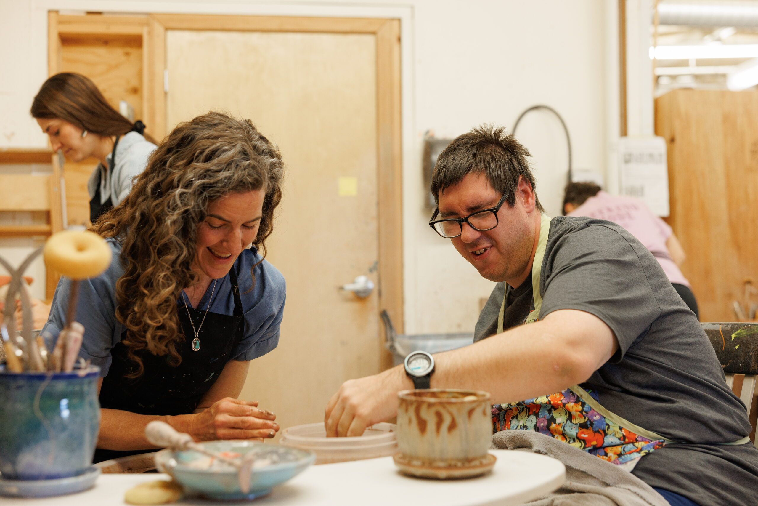 An instructor and an artist laugh together as they work clay on a pottery wheel, beside a glazed mug and bowl.