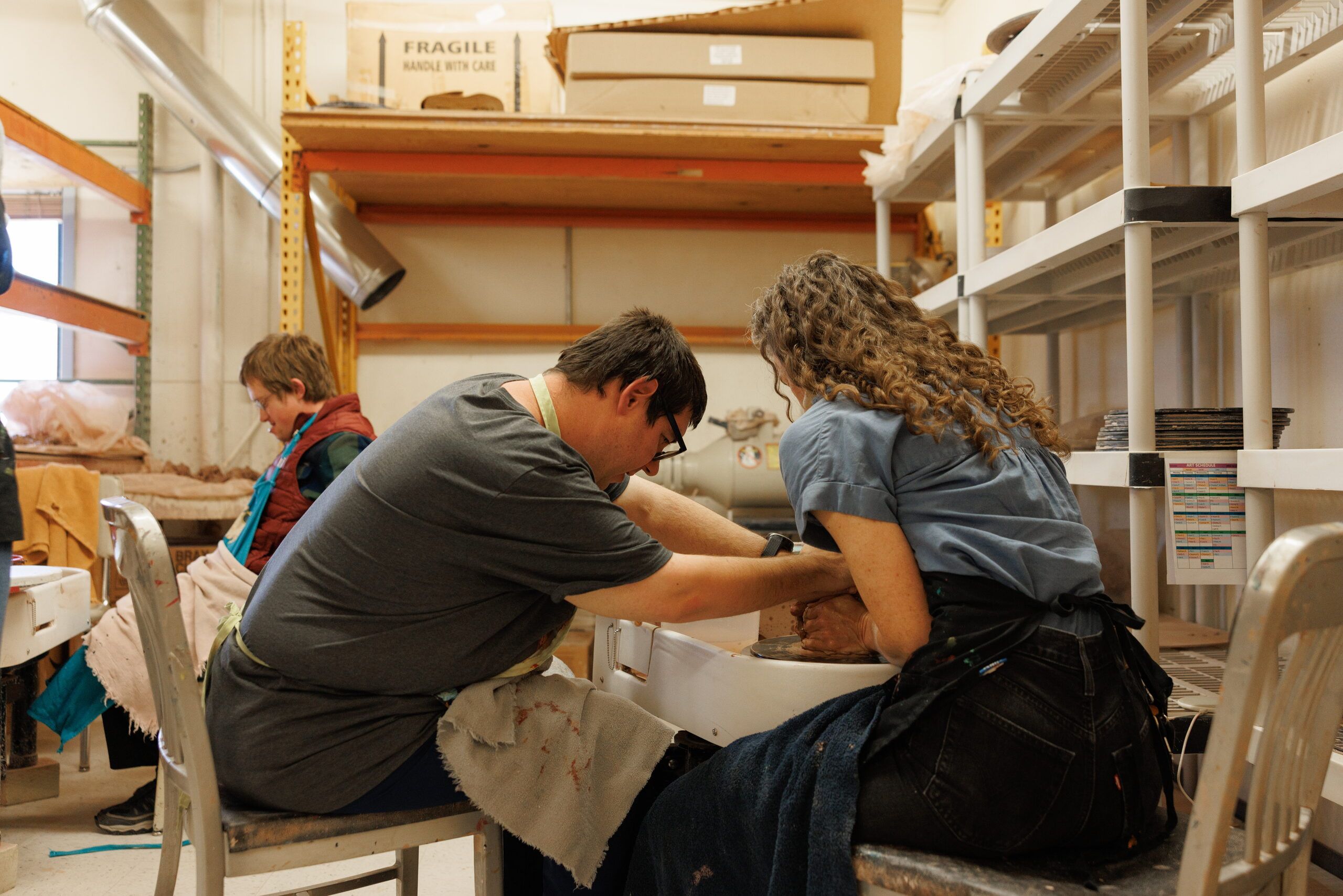 An instructor and an artist shape a piece of clay together on a pottery wheel, with studio shelves behind them.