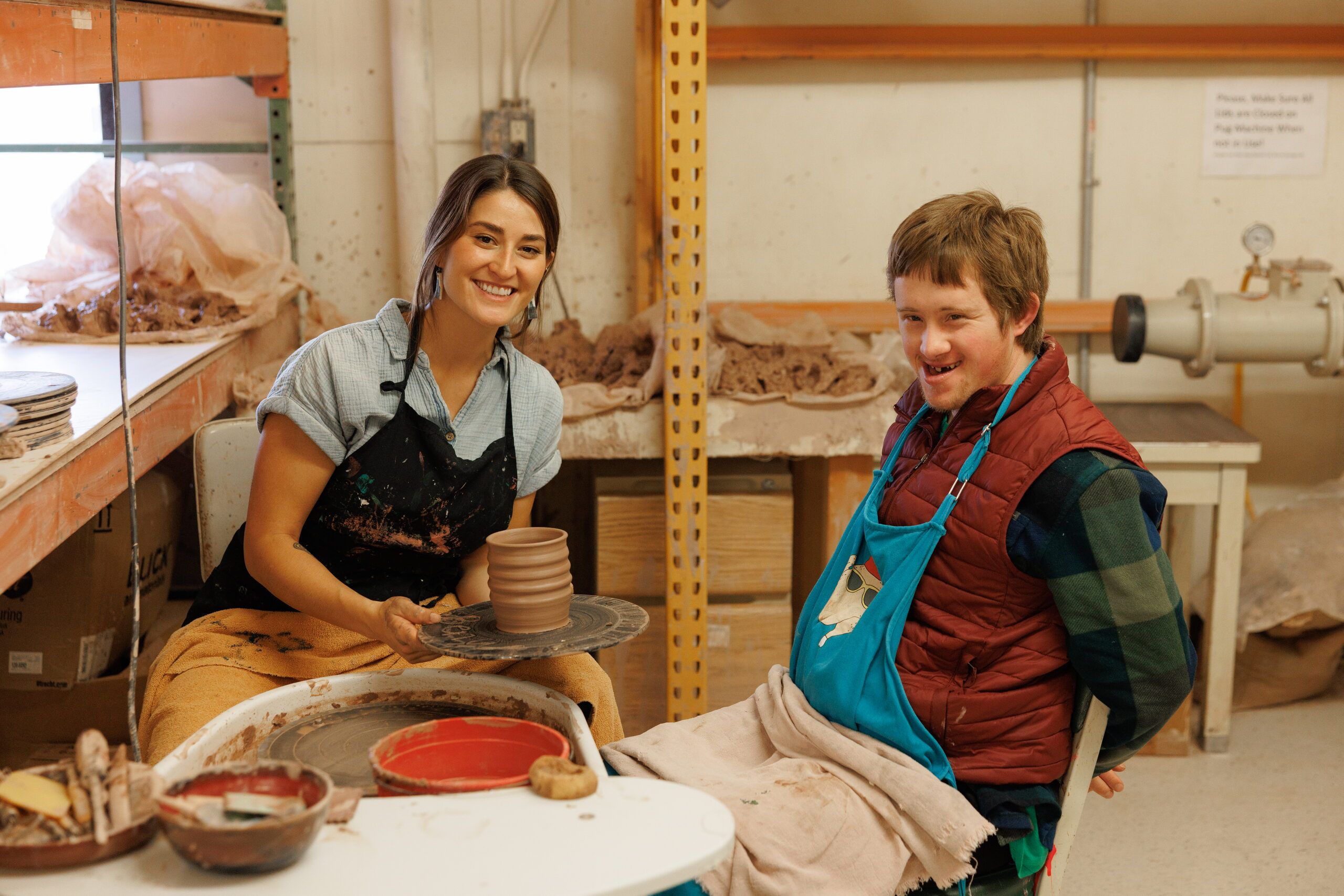 A smiling instructor holds a stack of freshly thrown clay pots on a wheel, next to a laughing artist in a teal apron.