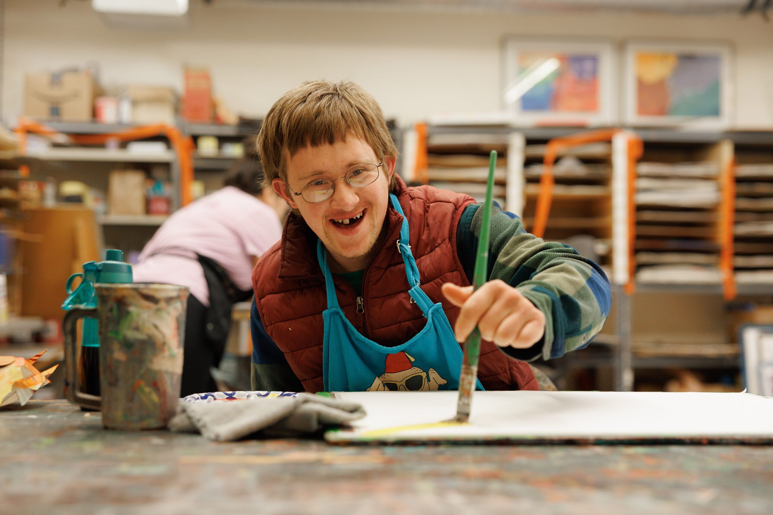 A smiling young artist in glasses and a teal apron brushes paint across a large sheet of paper in the 2D art studio.