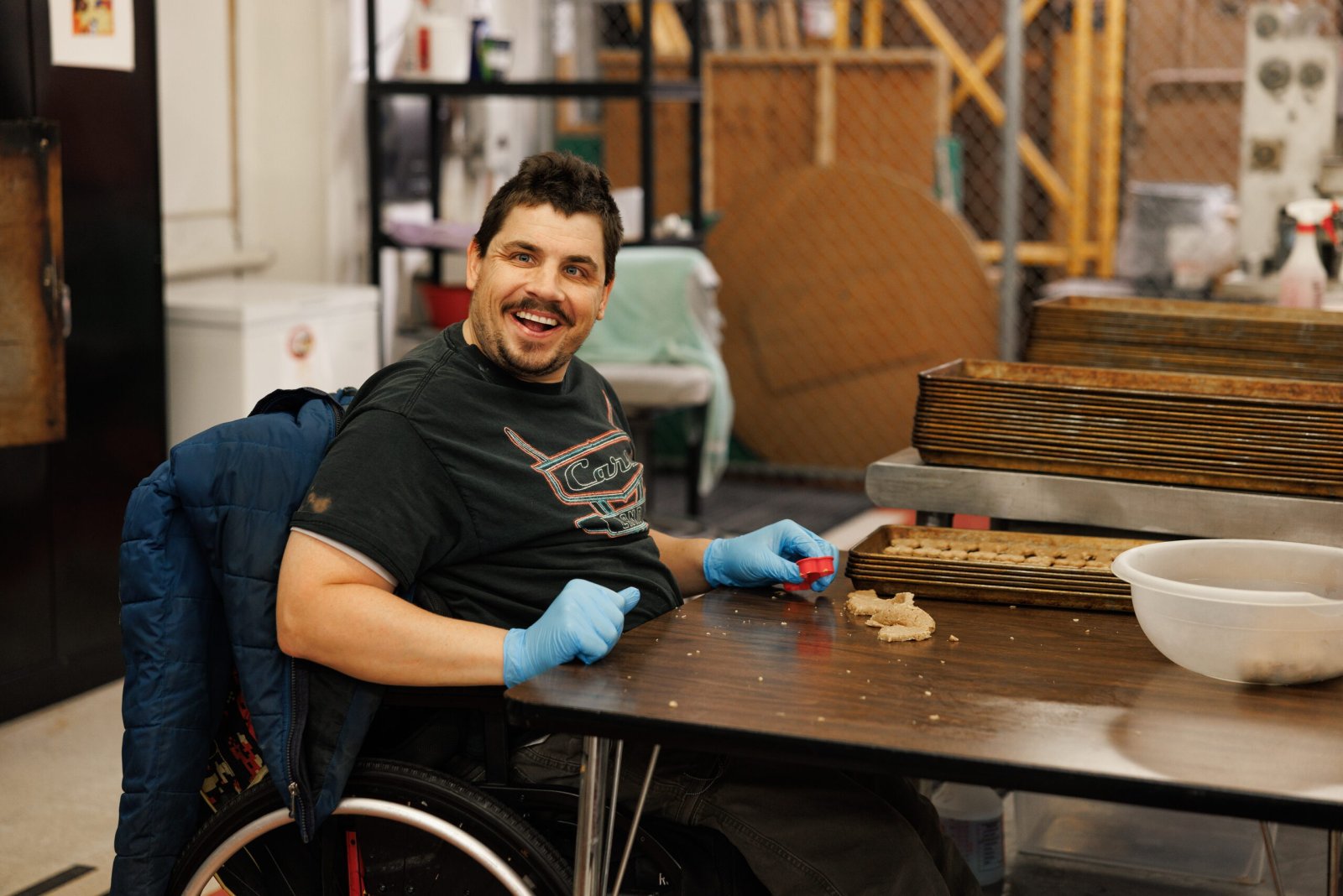 A smiling man in blue gloves shapes dog treats at a baking table.