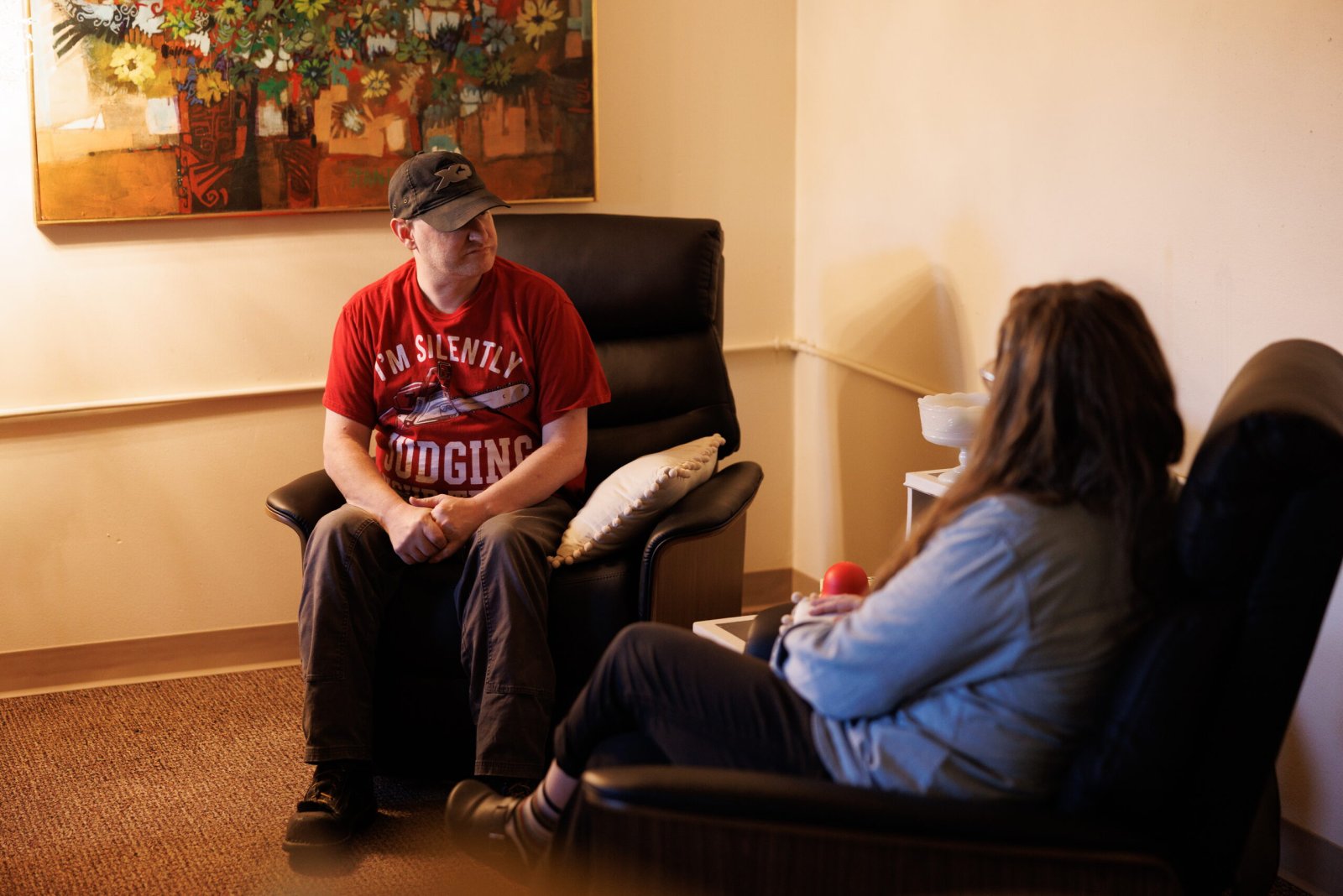 A man in a red shirt sits in an armchair, talking with a woman across from him.