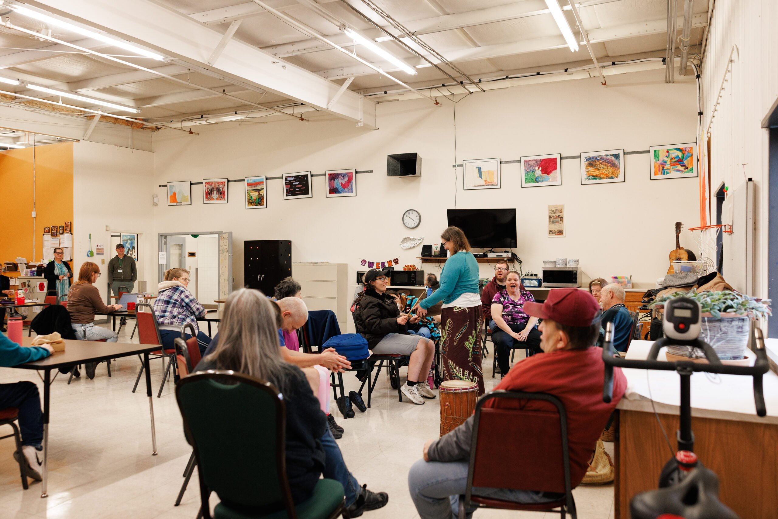 A large group sits in a circle with hand drums in a bright program room.