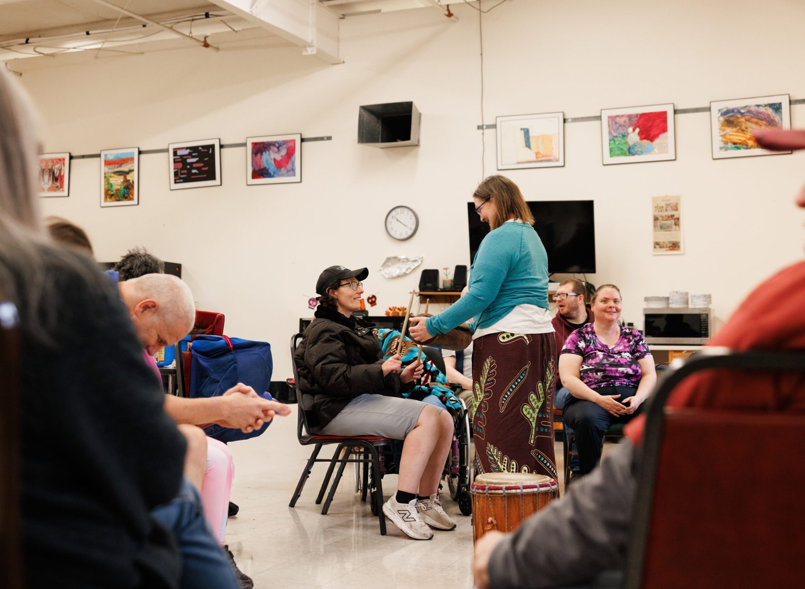 A woman with a hand drum leads a seated group in a bright program room.