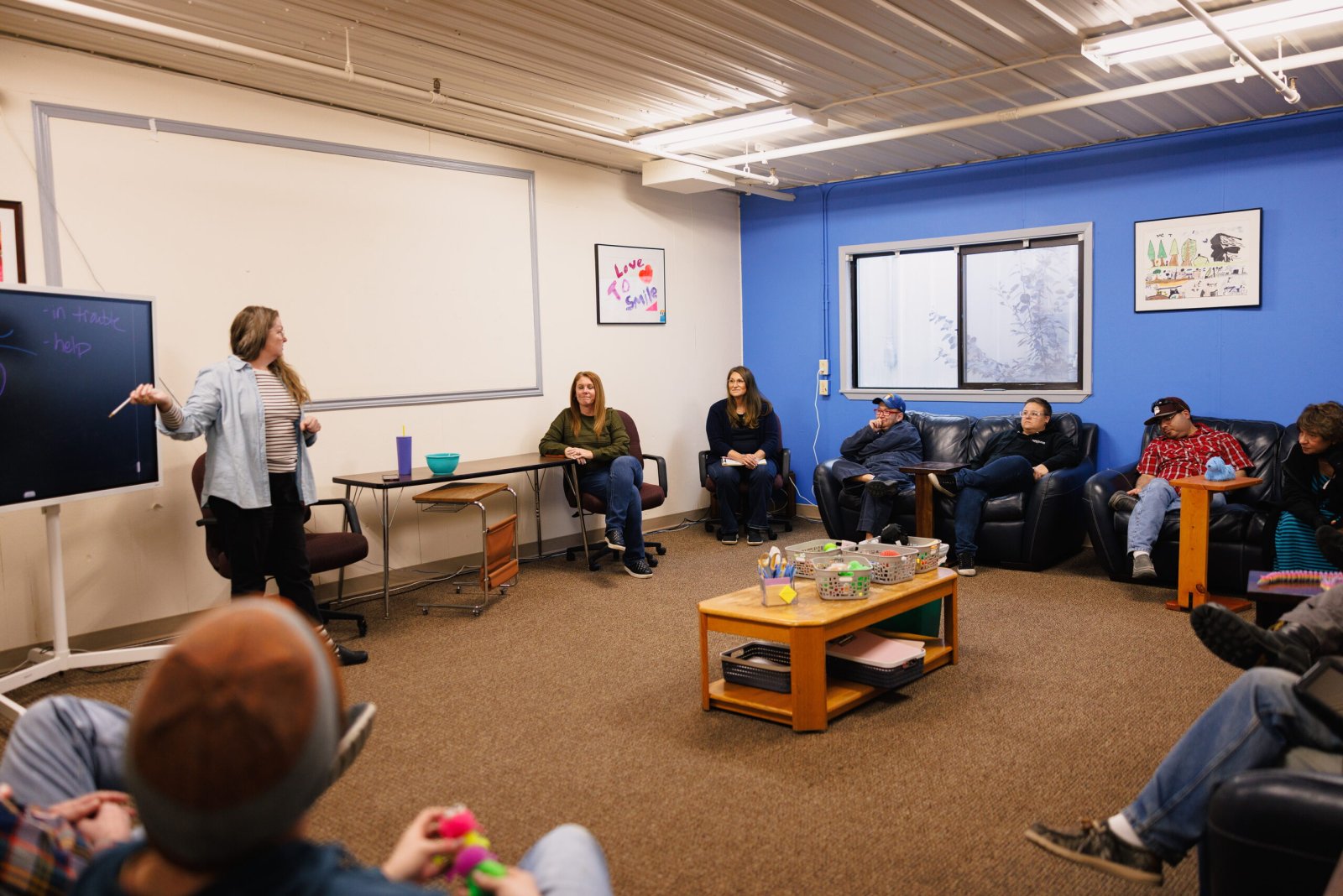 A woman at a whiteboard leads a group sitting on couches in a blue-walled room.