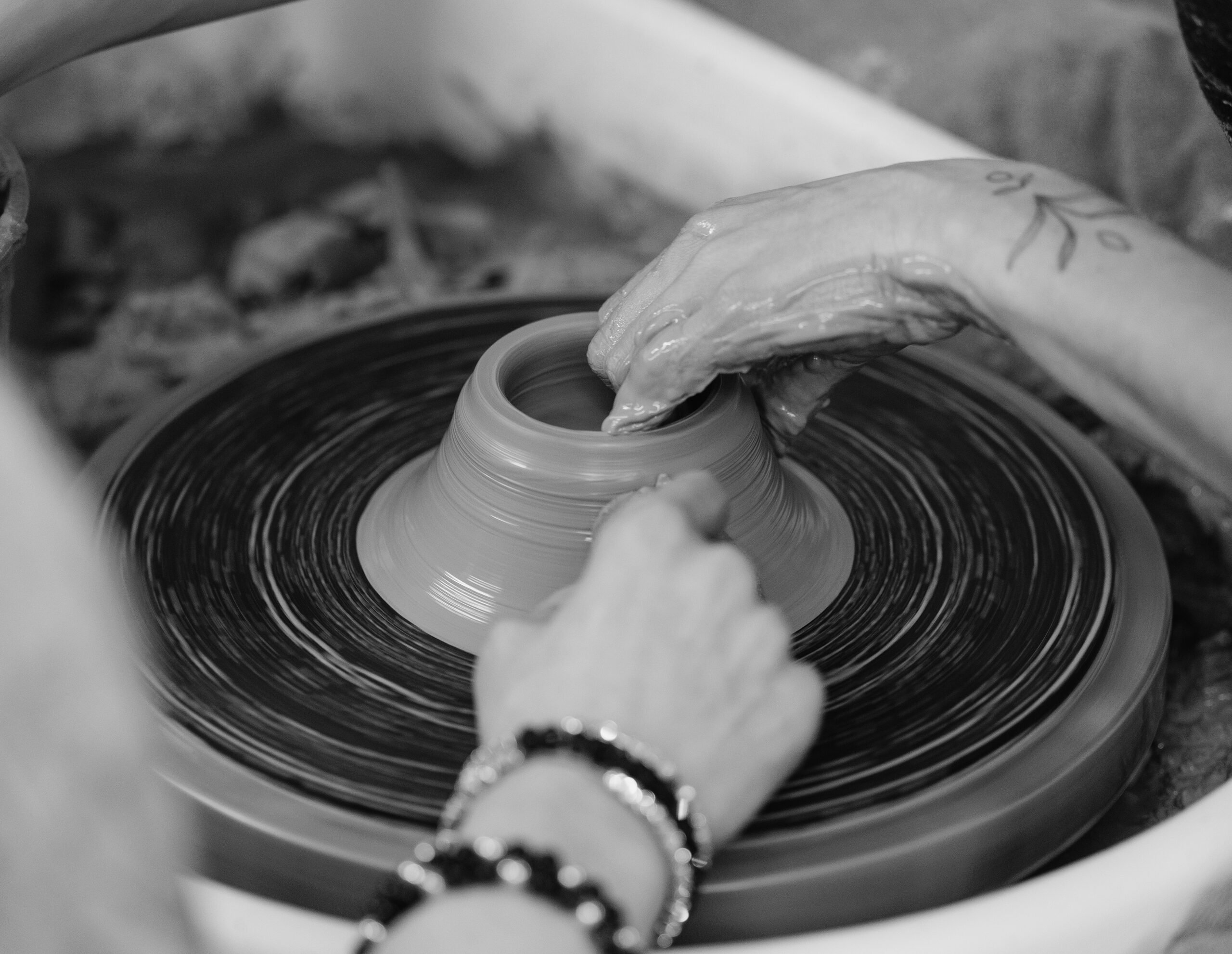 Hands shape a clay pot on a spinning pottery wheel, in black and white.