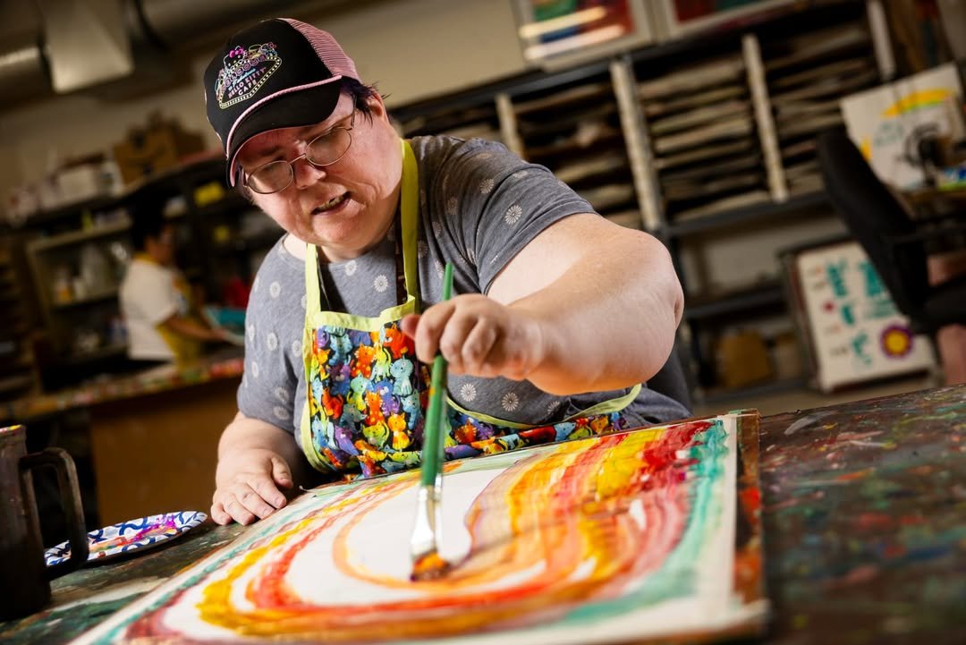 An artist in a cap and apron paints bright red, orange, and yellow swirls at a studio table.