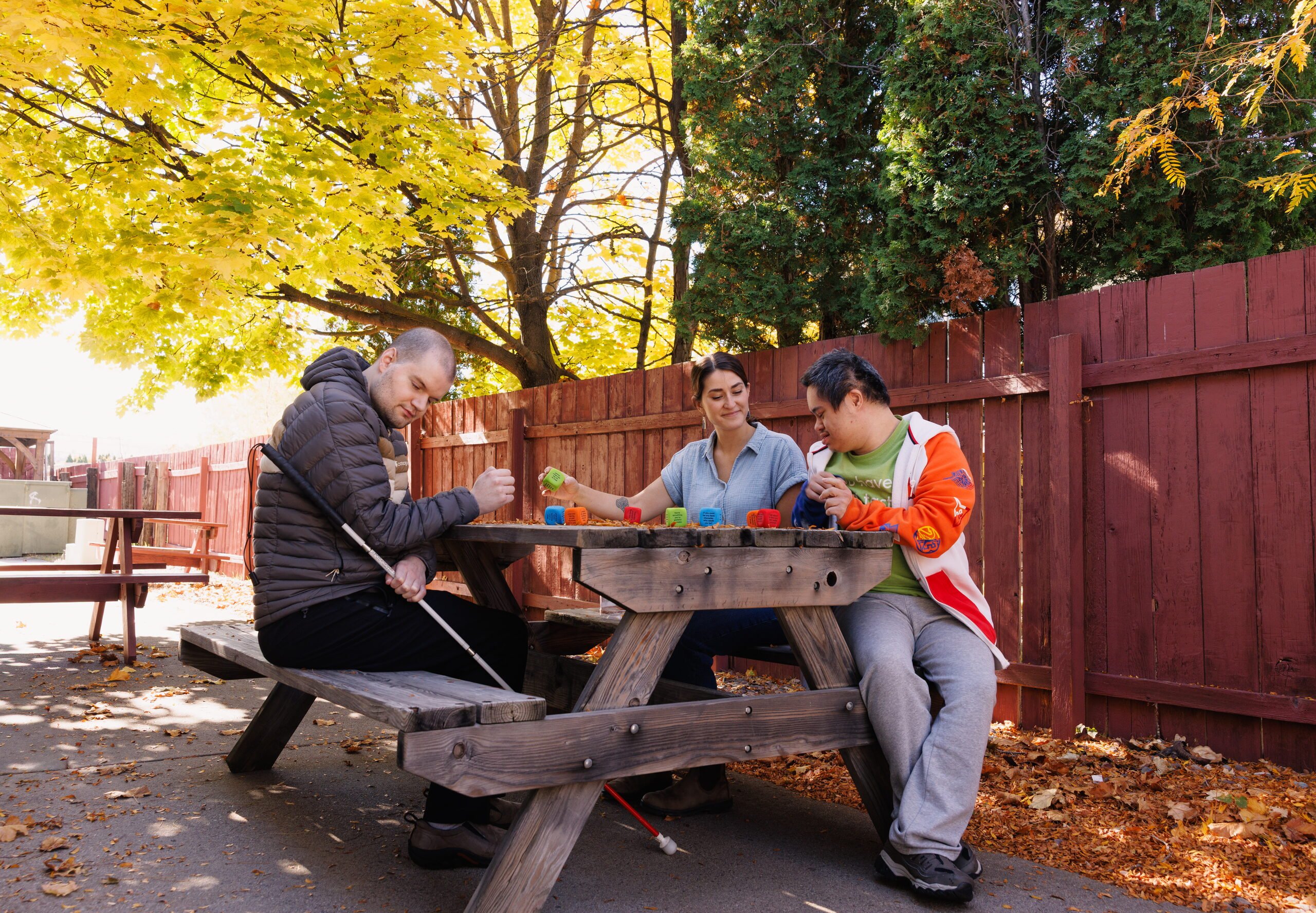 Three people sit at a picnic table under autumn trees, working on a colorful activity.